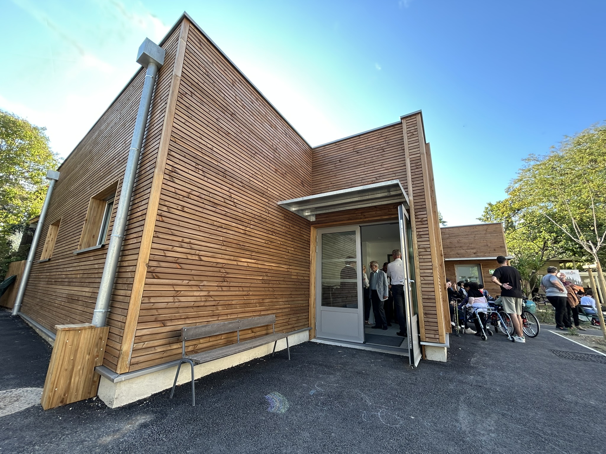 Entrée du nouveau bâtiment en bois de l'IEM La Cardabelle, récemment rénové et agrandi, avec des visiteurs et des résidents en fauteuil roulant profitant de l'inauguration.